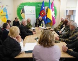 На світлині зафіксовано робочу зустріч групи людей за довгим дерев’яним столом у світлому офісному приміщенні, прикрашеному державною символікою та банером «Луганщина моя». Учасники, серед яких військові та цивільні, зосереджено слухають доповідача в центрі, що свідчить про обговорення важливих соціальних чи адміністративних питань регіону