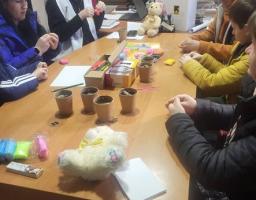 На світлині зображено групу людей, які сидять за довгим дерев'яним столом у кабінеті та займаються творчістю, ймовірно, ліпленням із пластиліну чи полімерної глини. На столі розкладено кольорові бруски матеріалу, стаканчики, олівці та м'які іграшки, а на задньому плані на стіні висить велика старовинна мапа Європи та календар