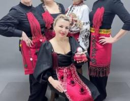 П'ять усміхнених жінок позують для групового фото в автентичному вбранні, що поєднує сучасний чорний одяг із традиційними українськими елементами — вишиванками та яскраво-червоними декоративними фартухами з вишивкою та бахромою