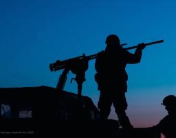 На світлині зображено темні силуети двох українських військових та великокаліберного кулемета на фоні глибокого синього неба в сутінках. Один із бійців стоїть біля турелі, перевіряючи готовність зброї, встановленої на військовому автомобілі бригади «Чорний ліс»