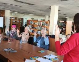 На світлині зображена група жінок, які сидять за великим столом у просторій бібліотеці та повторюють жести руками за жінкою в червоному светрі, що стоїть до камери спиною. Учасниці зустрічі, серед яких є люди старшого віку, зосереджено підняли руки вгору, а на фоні видно численні книжкові полиці, фліпчарт та навчальне приладдя на столі.