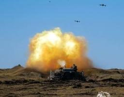 Динамічне фото бойової роботи української артилерії, де зафіксовано момент потужного вибуху та яскравого спалаху під час пострілу самохідної установки. Машина обладнана захисними протикумулятивними екранами («мангалом») та розташована на відкритій місцевості серед пагорбів під чистим блакитним небом.