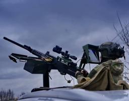 Військовослужбовець у повному бойовому спорядженні керує встановленим на пікапі великокаліберним кулеметом, оснащеним сучасним оптичним прицілом та виносним монітором для точного наведення на фоні похмурого неба.