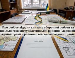 На великому дерев'яному столі в кабінеті Щастинської районної військової адміністрації розкладені топографічні карти, схеми евакуації та офіційні документи, обрамлені синьо-жовтими стрічками, тоді як на задньому плані через вікно видно захисні укріплення з мішків із піском