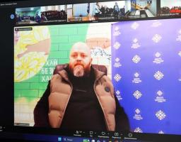 У центрі кадру — чоловік середнього віку з густою русявою бородою з сивиною. Він одягнений у чорну кофту та бежевий дутий жилет. Чоловік дивиться прямо в камеру.