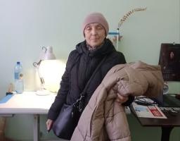 Одягнена в теплу чорну стьобану куртку та світло-рожеву (пудрову) в'язану шапку. Через плече у неї висить чорна сумка. Вона тримає в руках ще одну об'ємну довгу куртку бежевого або світло-коричневого кольору.