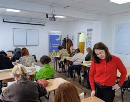 У кімнаті перебуває група жінок різного віку. Більшість із них сидять за партами, розставленими рядами, і зосереджено працюють із паперами — ймовірно, заповнюють анкети або виконують навчальні завдання.