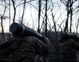 На фотографії в атмосфері сутінків або раннього світанку зображені двоє військових у камуфляжній формі, які йдуть крізь густий ліс із голими деревами. Кожен із них несе на плечі великий артилерійський снаряд або тубус від нього