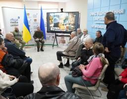 Фото зроблено з ракурсу ззаду глядачів, що сидять у колі. У центрі уваги — телевізійний екран із зображенням, що нагадує зону відчуження. Ліворуч стоять два прапори: державний прапор України та синій прапор із гербом. Спікер у військовій формі сидить із нотатником і звертається до аудиторії. Атмосфера офіційна та стримана.