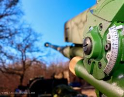 На зображенні представлено крупний план механізму прицілювання сучасної артилерійської установки зеленого кольору зі шкалою та цифрами, на тлі якої в розмитій перспективі видно ствол гармати, спрямований у безхмарне блакитне небо над лісистою місцевістю.