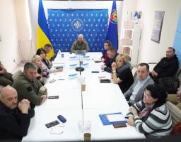 Група людей у цивільному та військовому одязі проводить офіційне засідання за великим білим столом у кабінеті, прикрашеному державною символікою України та інформаційними стендами.