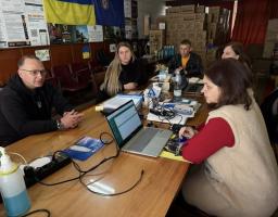 Група людей зосереджено працює за довгим дерев'яним столом, заставленим ноутбуками та документами, у приміщенні з українською символікою та численними коробками гуманітарної допомоги на задньому плані