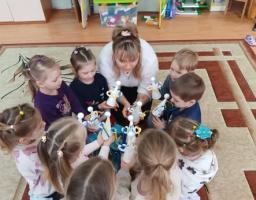  Вихователька та група з дев'яти маленьких дітей (хлопчиків та дівчаток) сиділи колом на килимі. Усі вони тримають у руках саморобні іграшки — білих паперових янголів з німбами.