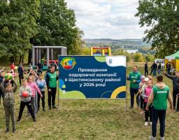 На лісовій галявині з надувними атракціонами та наметами група дітей і волонтерів у зелених жилетах проводить активні ігри з м’ячами та обручами навколо великого банера, що анонсує оздоровчу кампанію 2026 року в Щастинському районі.