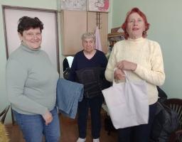 На знімку зображені три жінки старшого віку, які стоять у приміщенні. Вони дивляться в об'єктив і злегка посміхаються, створюючи атмосферу доброзичливості та взаємодопомоги