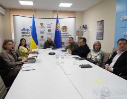 Група посадовців Луганської обласної державної адміністрації та представників медіацентру проводить робочу нараду за великим білим столом в офіційному кабінеті, прикрашеному державними прапорами та символікою адміністрації.