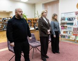 Троє людей — чоловік у чорному одязі та дві жінки у ділових костюмах — стоять у приміщенні бібліотеки поряд зі стелажами книг та кріслами, ймовірно, під час офіційного заходу чи хвилини мовчання.