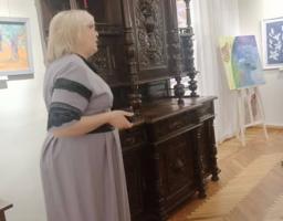 Жінка у довгій сірій сукні з мереживом проводить екскурсію у світлій музейній залі з паркетною підлогою, розповідаючи про масивний старовинний буфет із темного дерева, прикрашений складним художнім різьбленням.