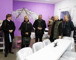 Група людей оглядає світлу кімнату з фіолетовими стінами, яка прикрашена до свята або тематичного заходу. У центрі — довгий білий стіл зі стільцями в білих чохлах. На задньому плані видно декоративне дерево з писанками та ангельські крила.