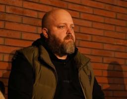 Портрет чоловіка з густою бородою, який сидить на фоні цегляної стіни. Чоловік одягнений у чорне худі та коричневий жилет, його погляд спрямований убік. Тепле вечірнє освітлення створює глибокі тіні.