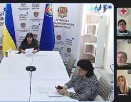 Світла конференц-зала з довгим білим столом. За столом сидять двоє жінок: одна на дальньому плані (спікерка, що вивчає документи), інша на передньому плані праворуч (у сорочці в клітинку).