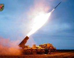 Фотографія бойової роботи реактивної системи залпового вогню (РСЗВ) на базі восьмиколісного вантажного шасі. Зафіксовано момент вильоту ракети з пускової установки, що супроводжується яскравим спалахом та вогняним слідом на фоні вечірнього неба.