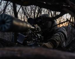 Український військовий у повному спорядженні та балаклаві веде спостереження через оптичний приціл гвинтівки з глушником, перебуваючи на замаскованій позиції серед густих гілок та лісової рослинності.