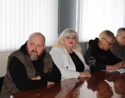 Четверо людей сидять за довгим полірованим столом під час зустрічі або наради. На задньому плані — вікна з білими вертикальними жалюзі. Учасники виглядають зосередженими, панує робоча атмосфера.