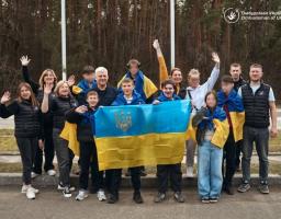 На знімку зображено близько 15 осіб — дітей різного віку та дорослих, які супроводжують їх. Вони стоять щільною групою на узбіччі дороги на фоні густого соснового лісу. Атмосфера сповнена радості, полегшення та патріотичного піднесення.