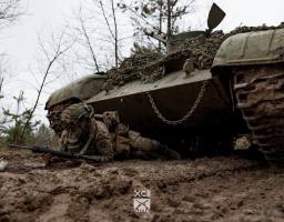 Український військовослужбовець у повному бойовому спорядженні та протигазі веде спостереження з автоматичною зброєю, перебуваючи в укритті безпосередньо під корпусом замаскованого бронеавтомобіля на ґрунтовій дорозі в лісистій місцевості.