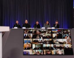 На фоні темних штор за довгим столом під час офіційного заходу сидять п'ятеро представників влади, серед яких у центрі Володимир Зеленський, а перед ними на великому похилому екрані відображається мозаїка з десятків вікон відеозв'язку з іншими учасниками конференції.