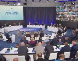 У великому сучасному залі під час заходу «Діалог громад з Парламентом» численні учасники в діловому вбранні розмістилися за напівкруглими столами навколо центральної восьмикутної трибуни, а на великих екранах відображаються назва панельної дискусії та десятки вікон із відеозв’язком дистанційних спікерів.