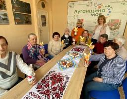 Група жінок старшого віку зібралася за довгим столом, застеленим вишитим рушником і прикрашеним крашанками, тримаючи в руках саморобні великодні сувеніри на тлі плаката з написом «Луганщина моя»