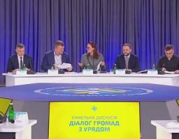 На тлі масивних синіх штор п'ятеро спікерів у діловому вбранні сидять за довгим білим столом із мікрофонами, проводячи панельну дискусію «Діалог громад з Урядом», назва якої яскраво світиться на жовтому екрані перед столом.
