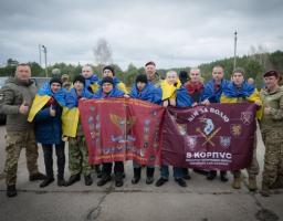 Група українських захисників та цивільних позує для спільного фото просто неба, тримаючи великі бордові прапори з символікою 8-го армійського корпусу та Десантно-штурмових військ, при цьому деякі учасники загорнуті в національні синьо-жовті стяги.
