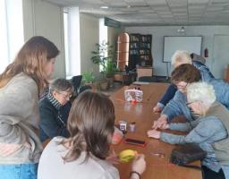 На фото зображено групу жінок різного віку, які зібралися навколо довгого дерев’яного столу в просторому приміщенні. Вони зосереджено займаються спільною справою, схожою на майстер-клас із рукоділля або волонтерську діяльність.