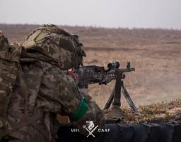 Військовослужбовець, знятий зі спини та збоку (у профіль). Він схилився над зброєю, зосереджено дивлячись у приціл.