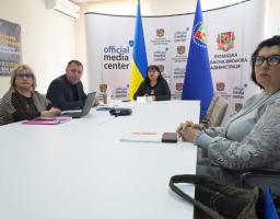 Четверо людей сидять за білим столом у приміщенні «Official Media Center» Луганської обласної військової адміністрації під час проведення брифінгу або робочої зустрічі на фоні державних прапорів.