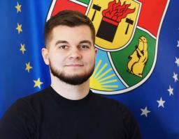 Портрет молодого чоловіка з темною бородою у чорній футболці на фоні синього прапора з жовтими зірками та великого кольорового герба Луганської області із зображенням бабака та сонця.