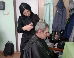 Жінка-перукар у чорному одязі стриже сивого чоловіка, накритого темним пеньюаром, у простому світлому приміщенні, де на фоні видно двері, вішалку з одягом та полиці з коробками