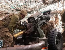 На фотографії український військовослужбовець у камуфляжній формі та шоломі, перебуваючи в укріпленій позиції під маскувальною сіткою, заряджає артилерійську гармату великим снарядом