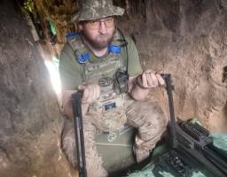На фото зображений бородатий український військовослужбовець в окулярах, камуфляжній панамі та плитоносці, який сидить у вузькому земляному окопі та тримає в руках деталі від розібраного великокаліберного кулемета, розкладеного перед ним на підстилці.