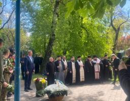 На фотографії шеренга православних священнослужителів у темному церковному вбранні та чоловік у синьому костюмі стоять біля великих кошиків із квітами під час офіційного пам'ятного заходу в оточенні густої зелені парку.