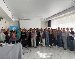 Група з понад тридцяти осіб різного віку позує для спільного фото у світлому конференц-залі перед білим екраном, де один чоловік сидить за столом із ноутбуком на передньому плані, а решта учасників стоїть щільною групою позаду нього.