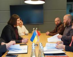 За довгим дерев'яним столом у сучасному офісному приміщенні з вимкненим екраном на стіні проходить офіційна зустріч шести осіб, де три жінки ліворуч та двоє чоловіків із жінкою праворуч обговорюють документи, сидячи навпроти маленьких настільних прапорців України та організації «Десяте квітня»