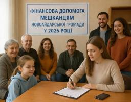 На світлині зображена група усміхнених людей різного віку в офісному приміщенні на фоні плаката «Фінансова допомога мешканцям Новоайдарської громади у 2026 році», де на передньому плані молода жінка підписує документи за столом поруч із дівчинкою.