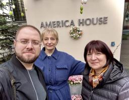 На фоні входу до будівлі з написом «AMERICA HOUSE», прикрашеного великоднім вінком та підвісними декоративними яйцями, троє усміхнених людей — чоловік в окулярах та чорній куртці і дві жінки в синьому пальті та темному пуховику — роблять спільне селфі.