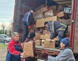 На вертикальній фотографії четверо людей розвантажують гуманітарну допомогу з великої червоної фури, заповненої картонними коробками, передаючи їх ланцюжком із кузова на землю на фоні похмурого неба та припаркованих автомобілів