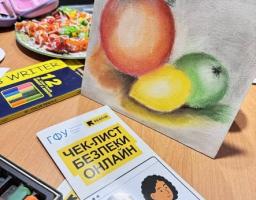 На дерев'яному столі поруч із художнім приладдям та тарілкою солодощів стоїть невелике полотно з натюрмортом, де зображені червоне яблуко, зелене яблуко та лимон, а на передньому плані лежить буклет із заголовком «ЧЕК-ЛИСТ БЕЗПЕКИ ОНЛАЙН», на якому намальовані дві дівчини з гаджетами.