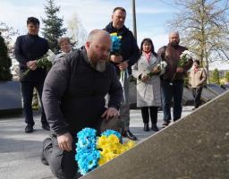 На передньому плані чоловік схилився на одне коліно біля кам'яної плити меморіалу, покладаючи квіти синьо-жовтих кольорів, тоді як позаду стоїть група людей із білими та синьо-жовтими букетами, вшановуючи пам'ять загиблих